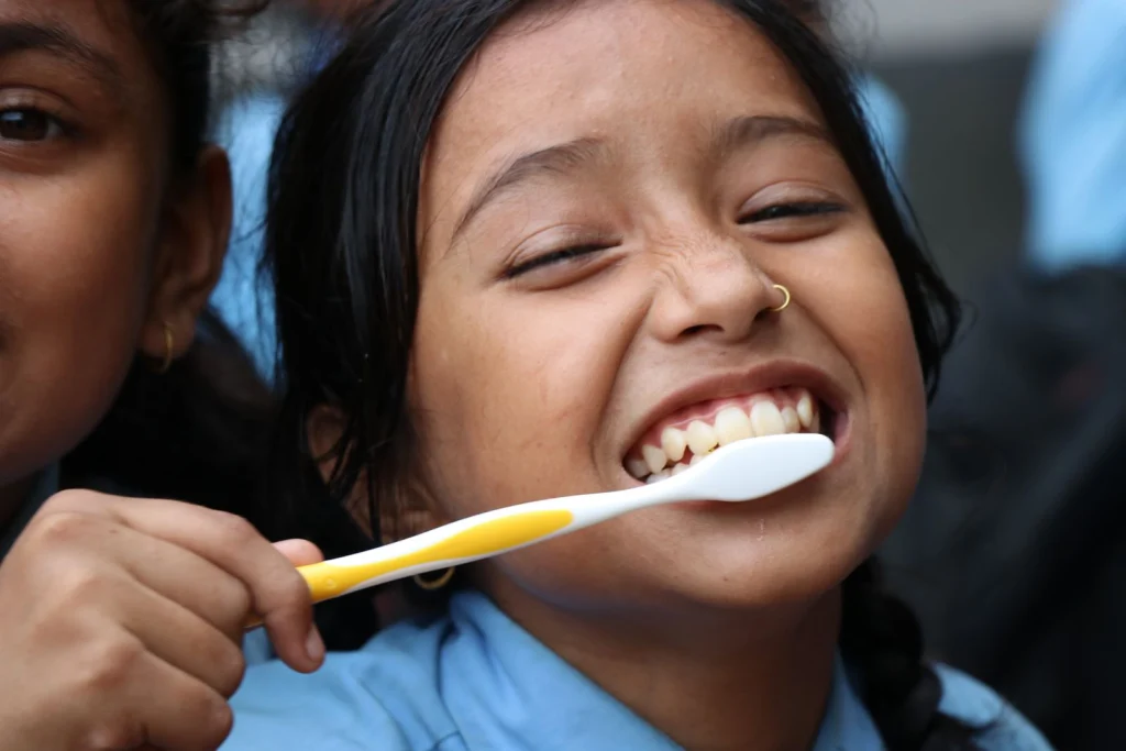 A cheerful child laughing while brushing her teeth with a yellow and white toothbrush.