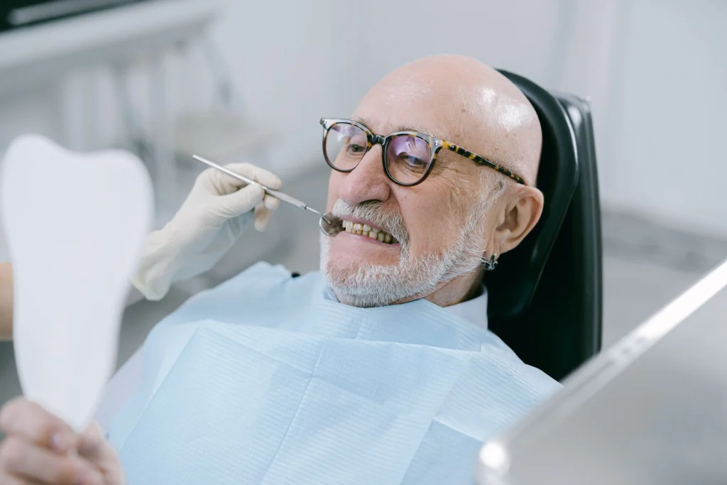 Elderly man at dental appointment, examining teeth with mirror.