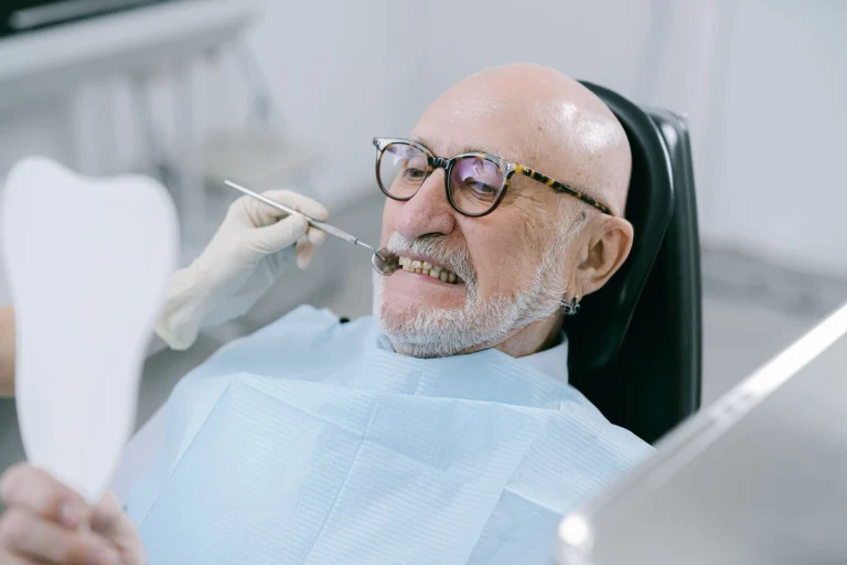Elderly man at dental appointment, examining teeth with mirror.
