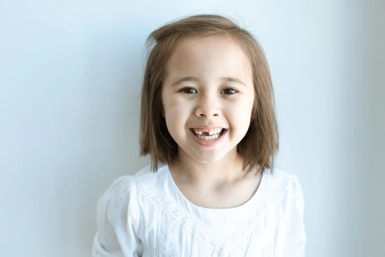 Cheerful young girl showing a missing tooth, wearing a white top against a plain background.