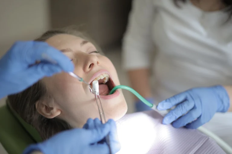 Unrecognizable crop dentists in latex gloves and uniform using professional medical equipment while treating teeth of young female patient sitting in chair with opened mouth and closed eyes in modern dental clinic