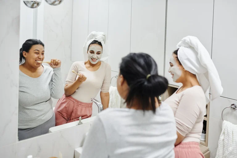 Two women enjoying skincare and oral hygiene routines with smiles in a modern bathroom mirror.