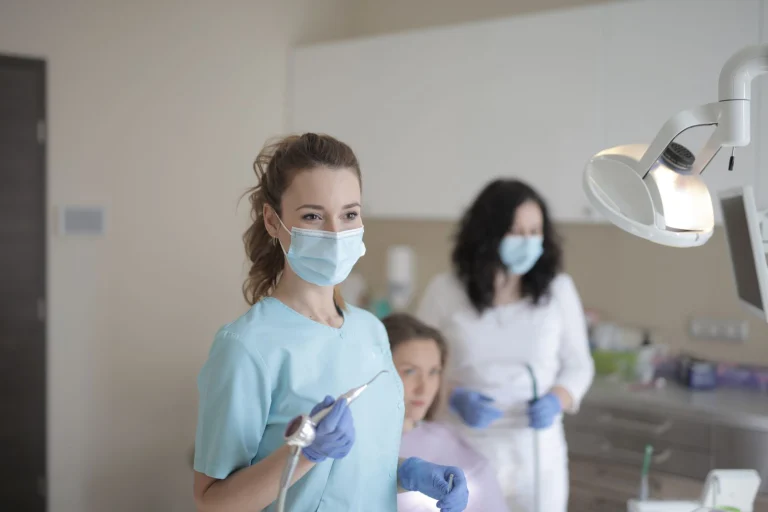 Young female dentist in medical mask and latex gloves using medical instrument while working with colleague during treatment of patient in modern dental room