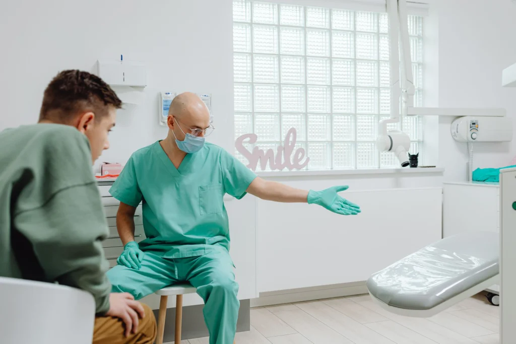 A dentist in green scrubs talks to a patient in a bright, modern dental clinic.