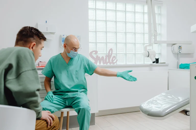 A dentist in green scrubs talks to a patient in a bright, modern dental clinic.