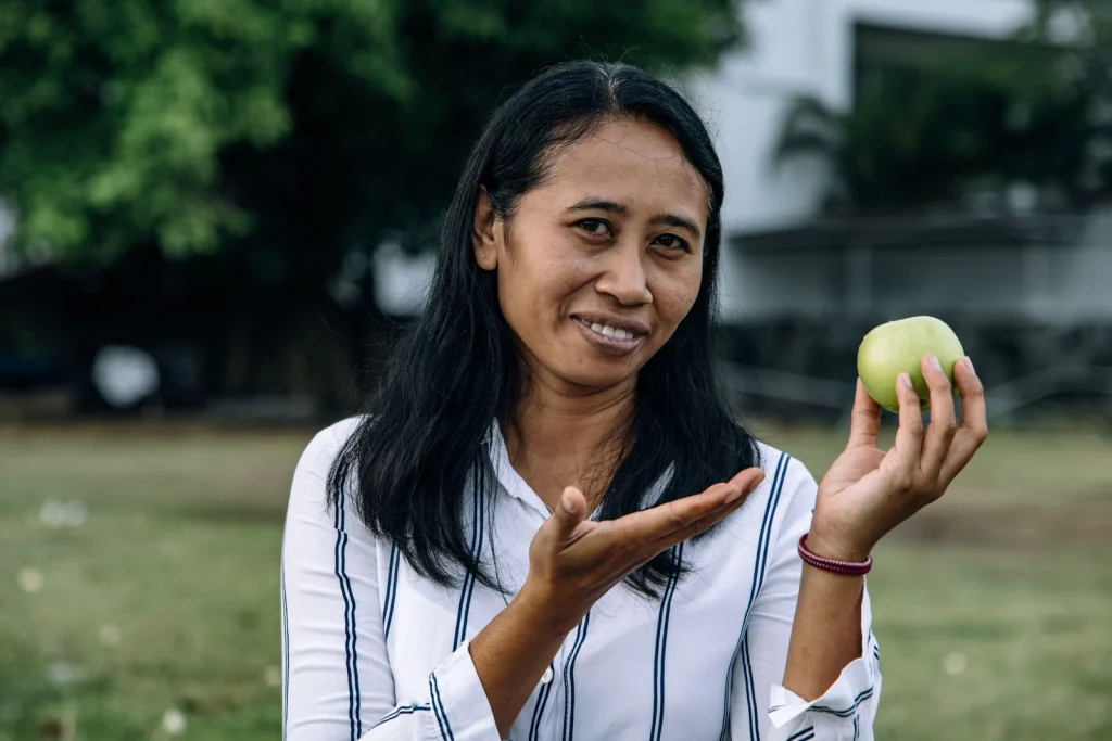 Smiling Asian woman presenting a green apple in an outdoor setting.