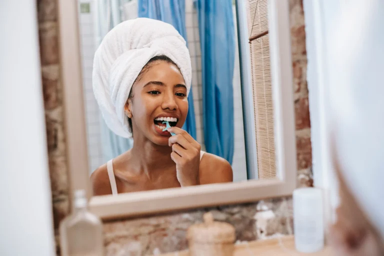 Reflection of young African American female with white towel on head looking in mirror while cleaning teeth with dental flosser