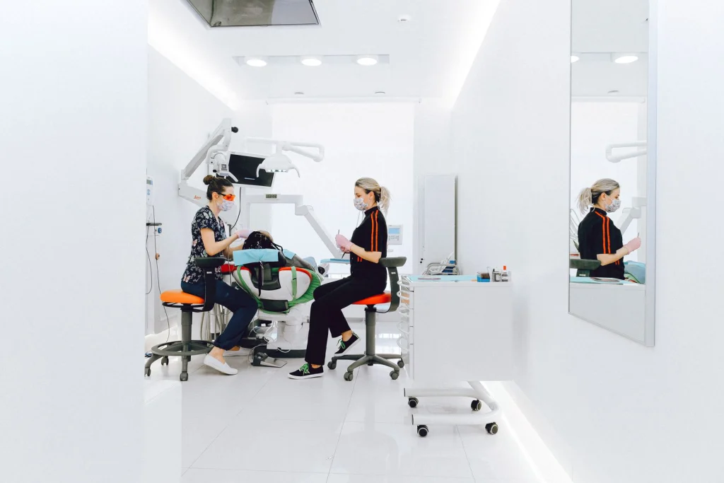 Side view of stomatologist and nurse in face mask checking up patients teeth while sitting on chairs near medical equipment in light doctor office