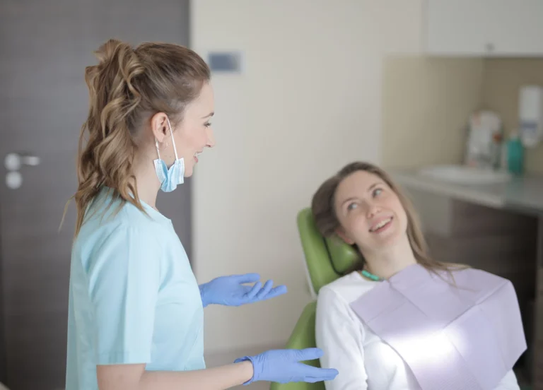 Positive young female dentist in latex gloves and uniform talking with young patient while sitting near chair and preparing for treatment in modern dental room in hospital