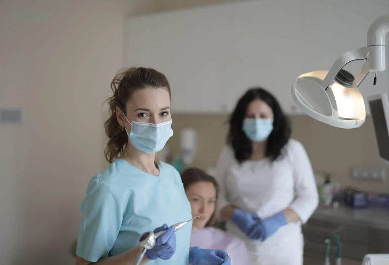 Female dentists in medical masks and latex gloves wearing uniform and treating teeth of patient while standing in modern dental room and looking at camera