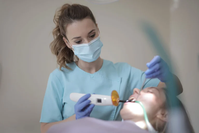 Side view of young female dentist in medical mask and latex gloves wearing blue uniform using modern medical equipment while treating teeth of patient in modern dental room in clinic