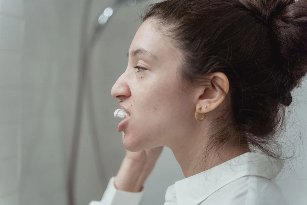 Side view of a woman with hair bun brushing teeth as part of her hygiene routine indoors.