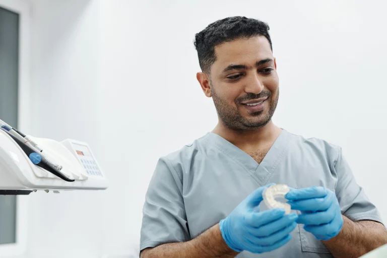 A dentist in scrubs and gloves examines a dental mold in a bright clinic setting.